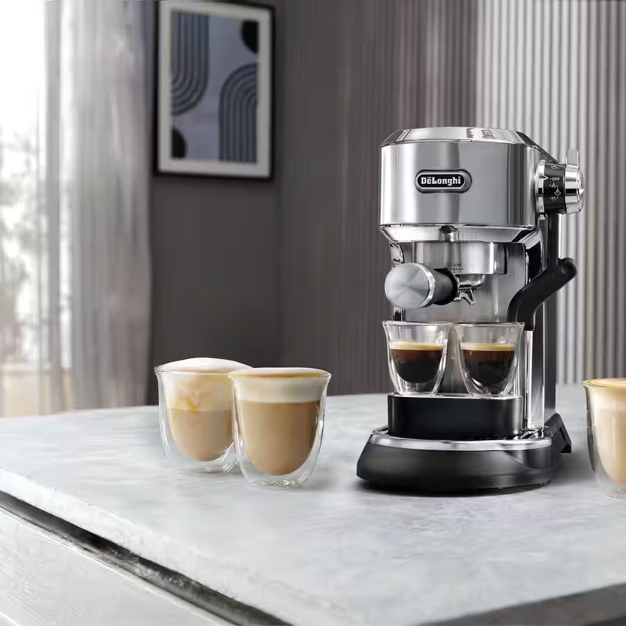 A De'Longhi espresso machine in silver and black sits on a light marble countertop, with two small espresso cups and two double-walled latte cups nearby in a modern kitchen setting.