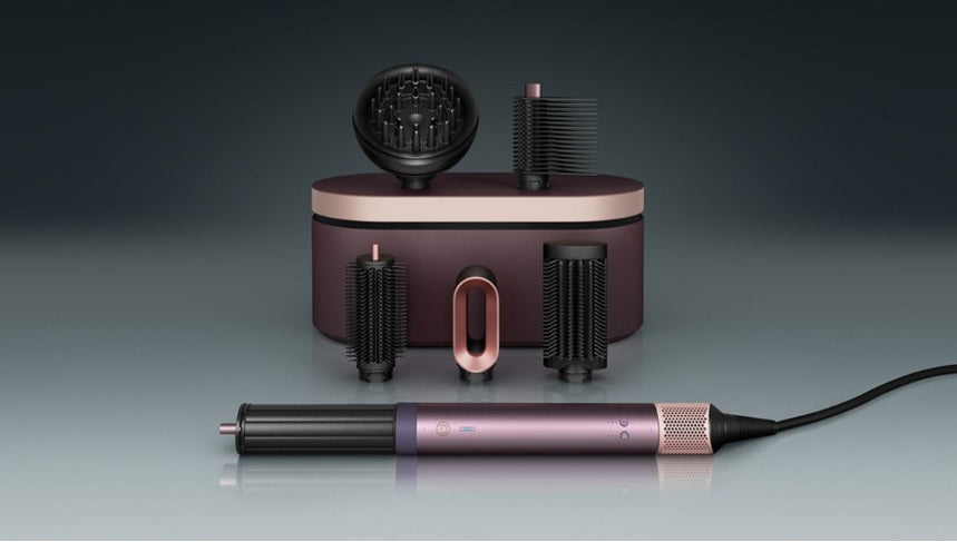 A hair styling tool set with a metallic purple and rose gold cylindrical wand, several black brush attachments, and a matching charging case on a dark gray gradient background.