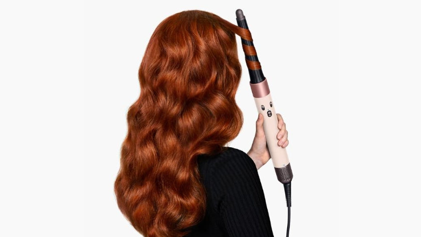 A woman with long, wavy red hair is shown from the back, holding a curling iron that is curling her hair, against a plain white background.