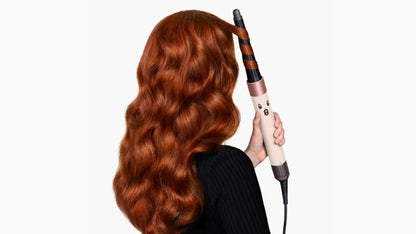 A woman with long, wavy red hair is shown from the back, holding a curling iron that is curling her hair, against a plain white background.