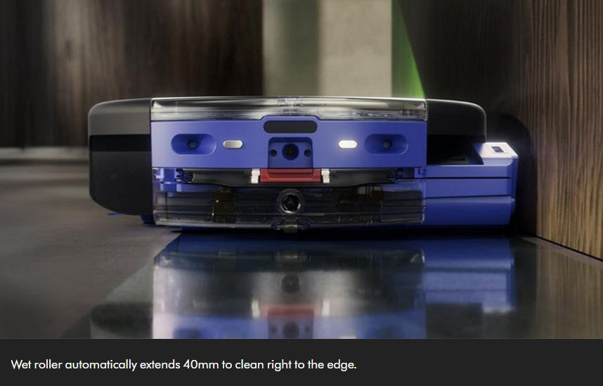 A blue and black robotic vacuum cleaner shown from the back, positioned on a glossy dark surface against a wood-paneled wall. A text overlay at the bottom reads, 'Wet roller automatically extends 40mm to clean right to the edge.