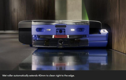 A blue and black robotic vacuum cleaner shown from the back, positioned on a glossy dark surface against a wood-paneled wall. A text overlay at the bottom reads, 'Wet roller automatically extends 40mm to clean right to the edge.