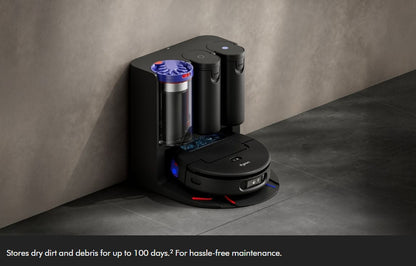 A black robotic vacuum cleaner docked at a matching black charging and cleaning station against a textured gray wall and dark floor. The station includes a transparent dust container with blue lighting accents.