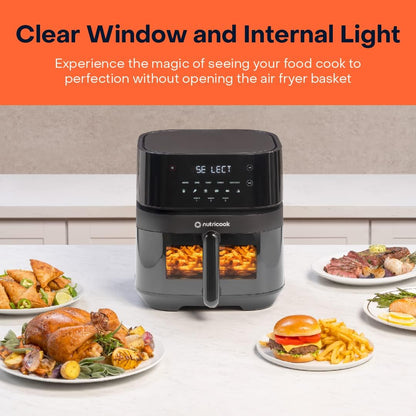 Black air fryer with clear window displaying food, surrounded by plates of cooked meals on a kitchen counter.